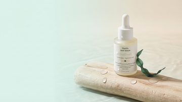Maren Dew Serum hydrating facial serum in frosted glass dropper bottle placed on natural driftwood with fresh water droplets and ocean kelp, minimal coastal aesthetic with soft green and sand tones